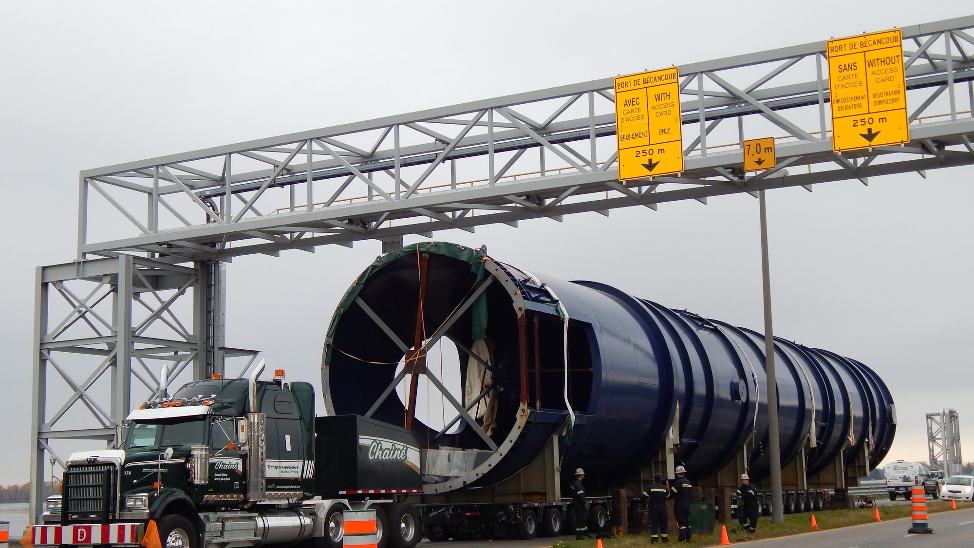 Le convoi Chainé passe sous le portique d’acier du Port de Bécancour : le tracteur vert devant, les segments bleus de la tour de refroidissement sur le transporteur, des cônes orange sur la chaussée