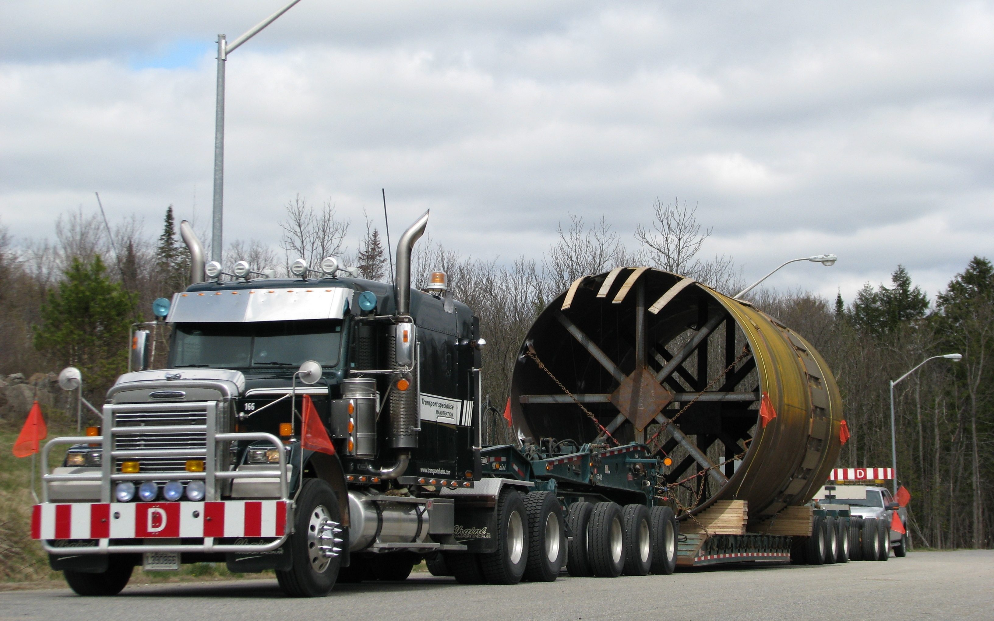 Le nez d’un tracteur Western Star vert Chainé avec sa plaque « D », un tambour d’acier de cinq mètres sur le fardier derrière lui, sur une route de campagne