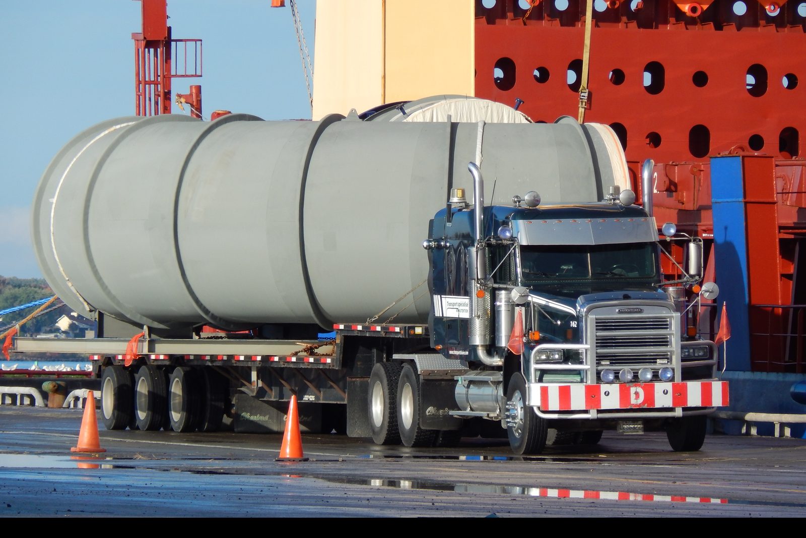 Un long réservoir gris sur une remorque Chainé au Port de Bécancour, la coque orange d’un navire derrière